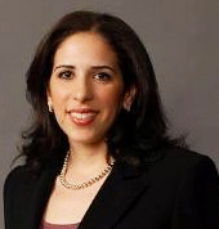 Photograph of Jill Keblawi, presenting as a female with light brown skin and long, dark brown hair. She is smiling.