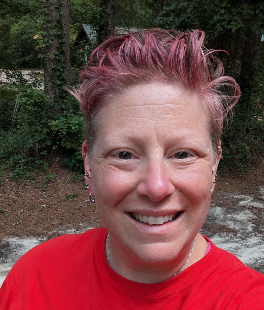 A female-presenting woman with light hair dyed red and smiling at the camera.
